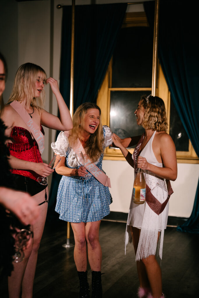 A group of women are celebrating a hens party in Sydney CBD at The Golden Era studio. They are doing a pole dancing and burlesque lesson and having so much fun, all dressed up and smiling.