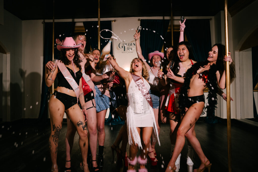 A group of women are celebrating a hens party in Sydney CBD at The Golden Era studio. They are doing a pole dancing and burlesque lesson and having so much fun, all dressed up and smiling.